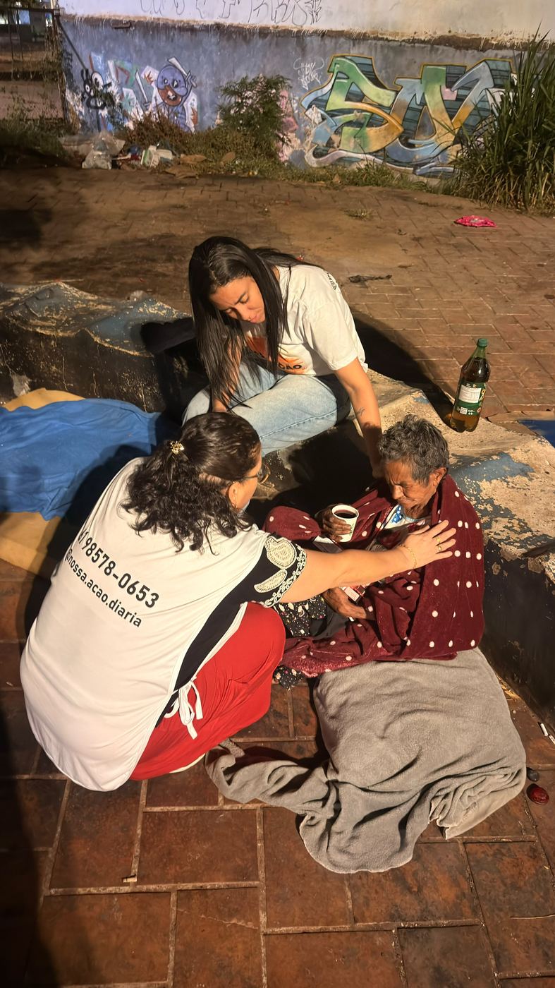 Voluntárias cuidando de uma senhora sentada na calçada, enrolada num cobertor