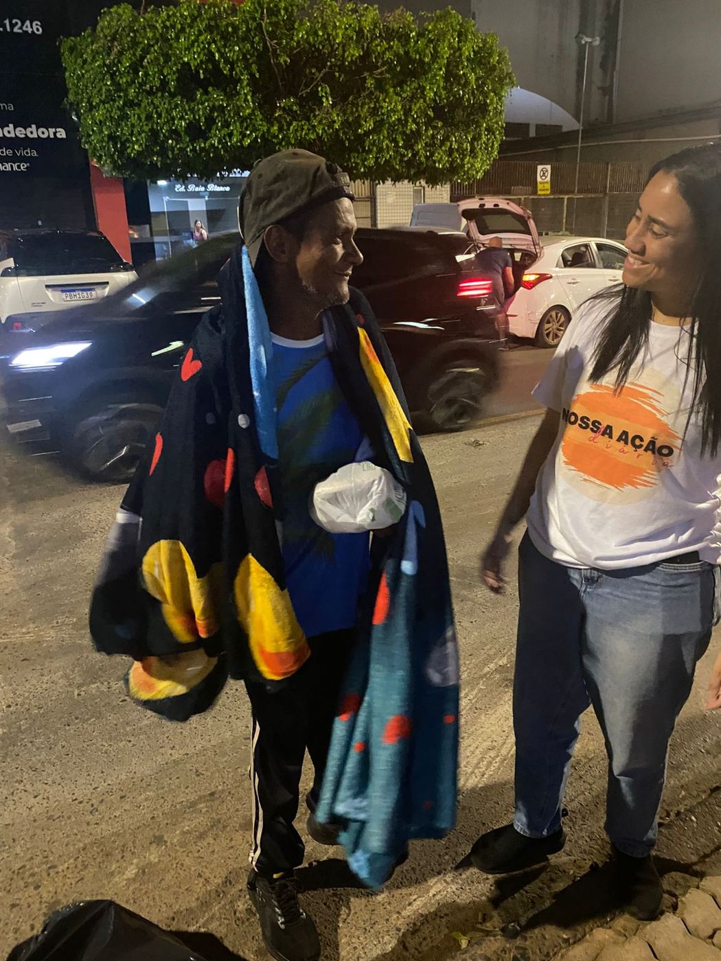 Homem recebendo cobertores de uma voluntária na rua, à noite